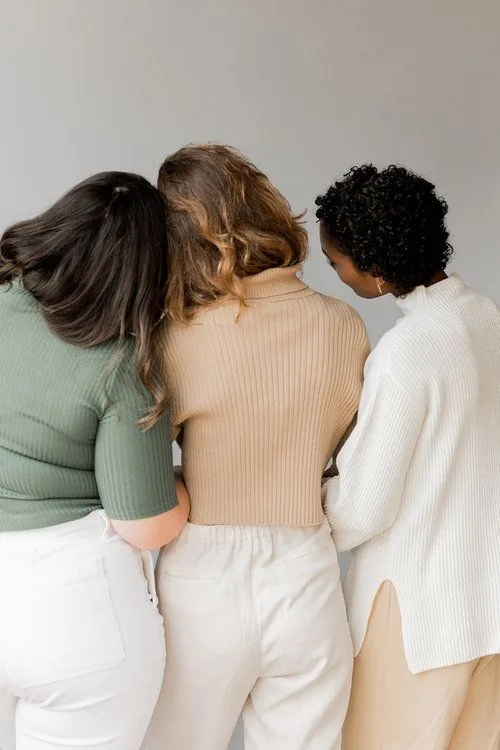 Três mulheres lado a lado, de costas, apoiadas umas nas outras