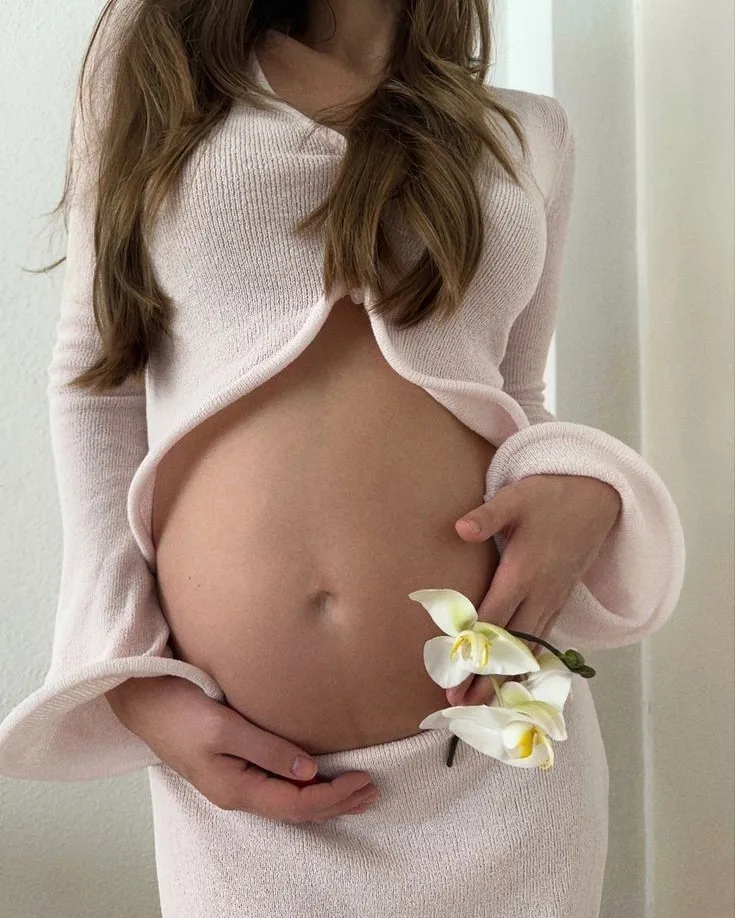 Gestante de suéter rosa claro segurando uma orquídea branca junto à barriga